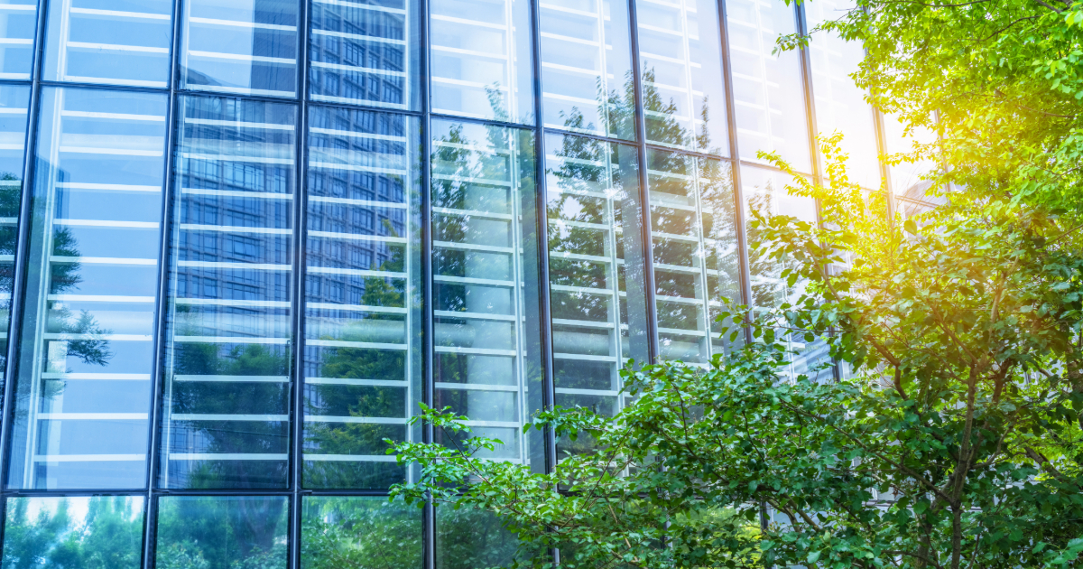 Exterior view of a commercial building with a large glass curtain wall facade reflecting trees and sky on a sunny day