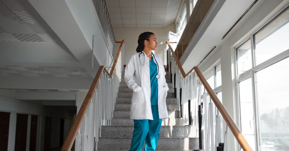 Healthcare worker in scrubs and white coat standing in a sunlit hospital corridor beside large windows