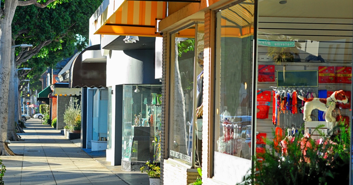 Row of retail storefronts with large glass display windows along a tree-lined commercial sidewalk.